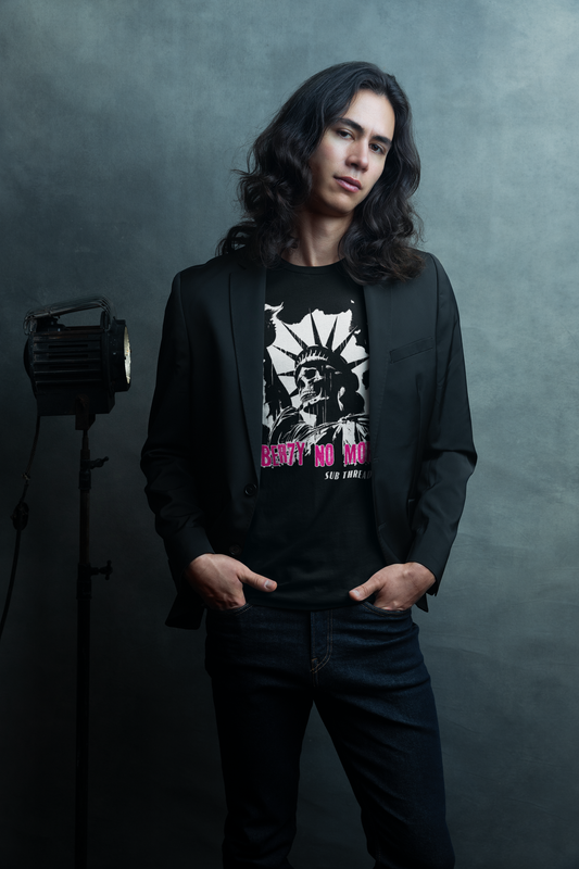 Young man in a studio for an editorial shoot, wearing a dark jacket and jeans. His black organic cotton Sub Threads shirt has a black and white design of the statue of liberty shown with a skull instead of her face. Magenta lettering states 'liberty no more'. He looks cool in his Sub Threads T-shirt pondering how the sustainable and eco-friendly production and fulfilment of Sub Threads products can help him look this cool while he's doing his bit to help the planet.